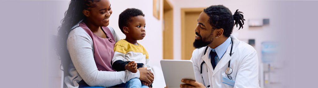 A doctor kneeling and holding a device, talking to a child who is sitting on an adult woman's lap.